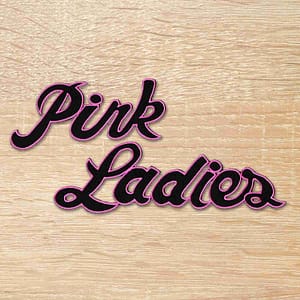 The word Pink Ladies Patch 50's Iron on Patch is written on a wooden surface.